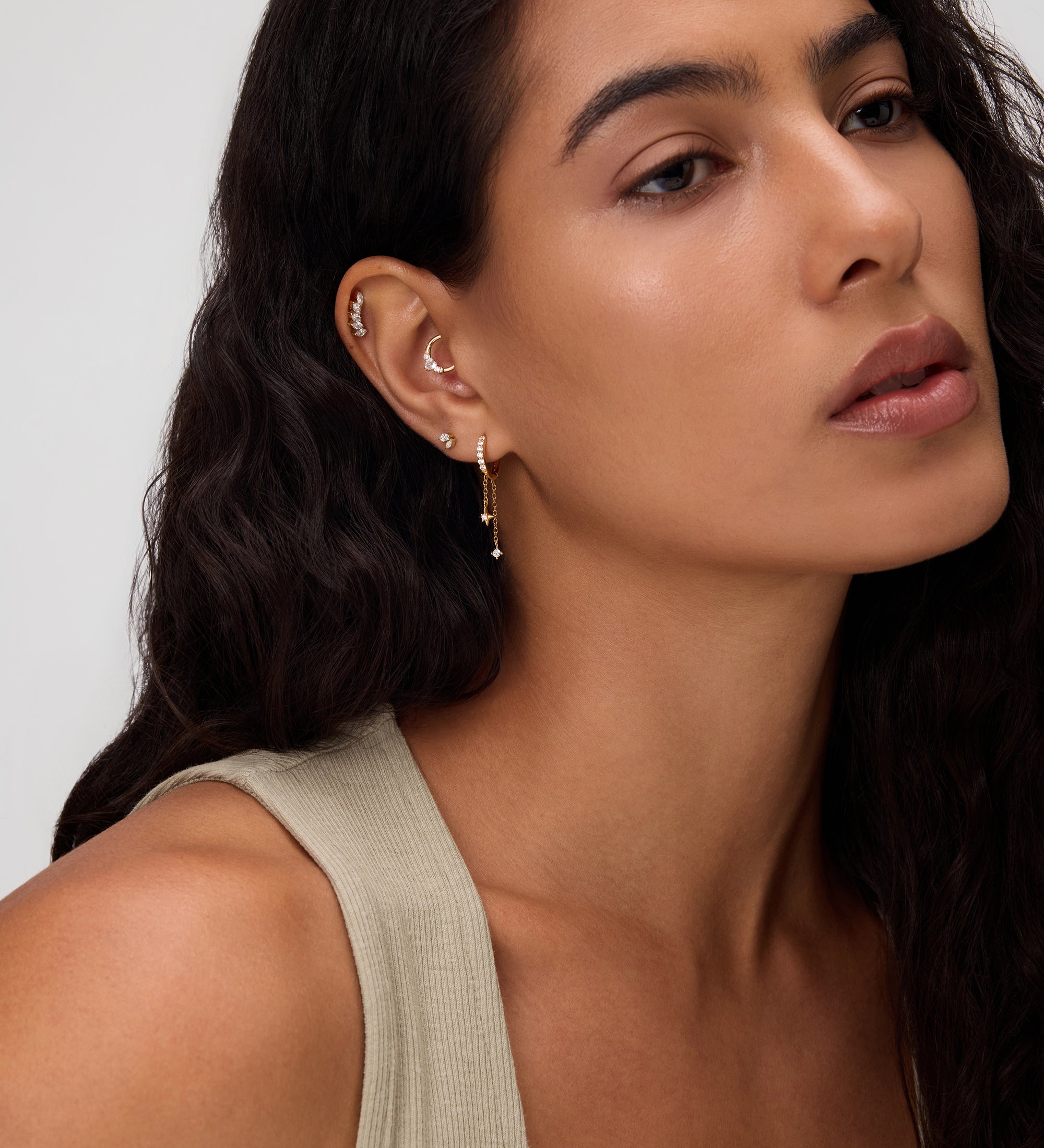 A woman with long, wavy dark hair wears a beige sleeveless top and 18k gold plated Star Double Chain Dangle Huggies in Gold with cubic zirconia. She looks calm, with natural makeup against a neutral background.