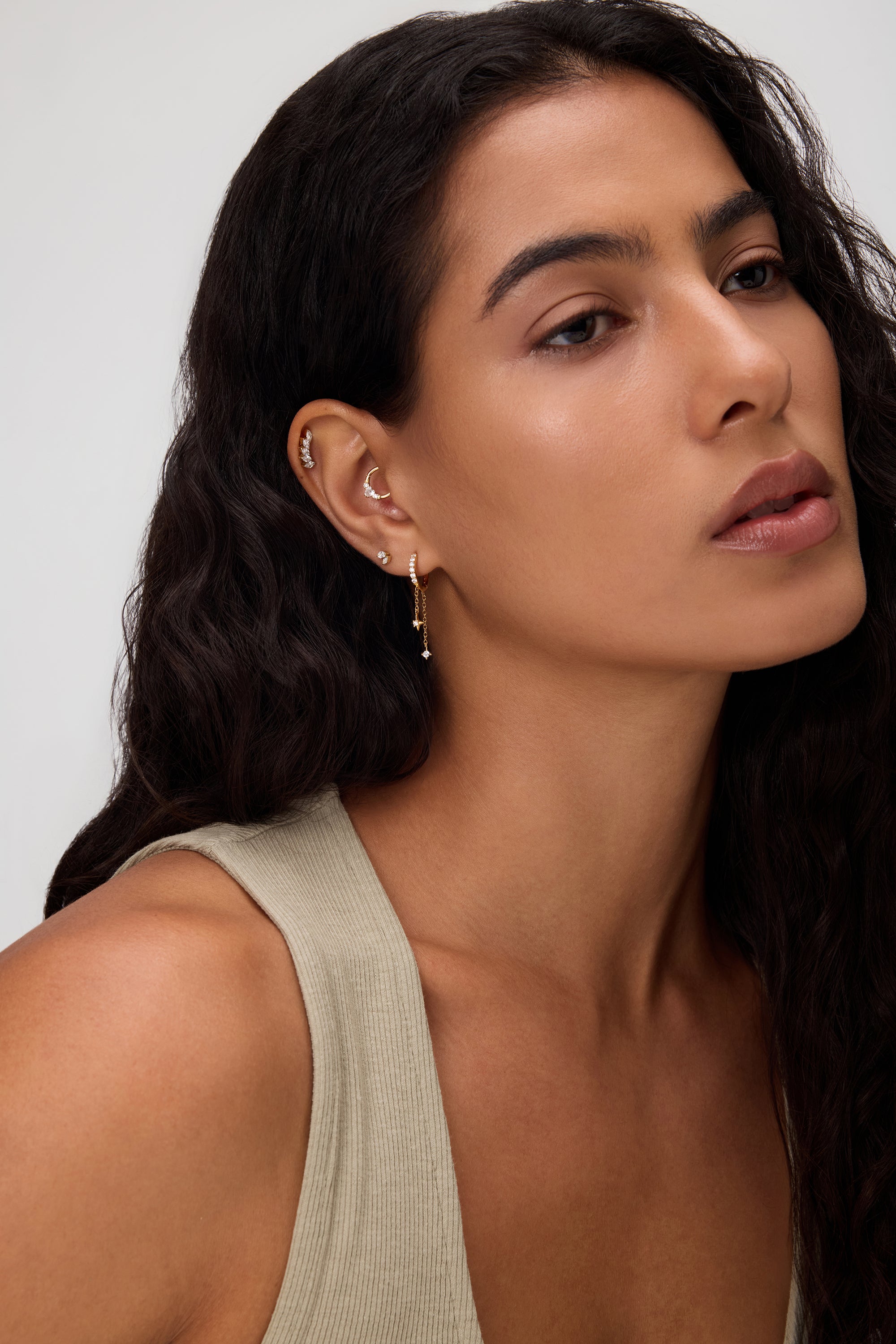 A woman with long, wavy dark hair wears a beige sleeveless top and 18k gold plated Star Double Chain Dangle Huggies in Gold with cubic zirconia. She looks calm, with natural makeup against a neutral background.