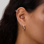 Close-up of a person's ear adorned with multiple earrings, including the Nori Twisted Hoop Earrings in Gold on the lobe. The person has long, dark hair and light brown skin.