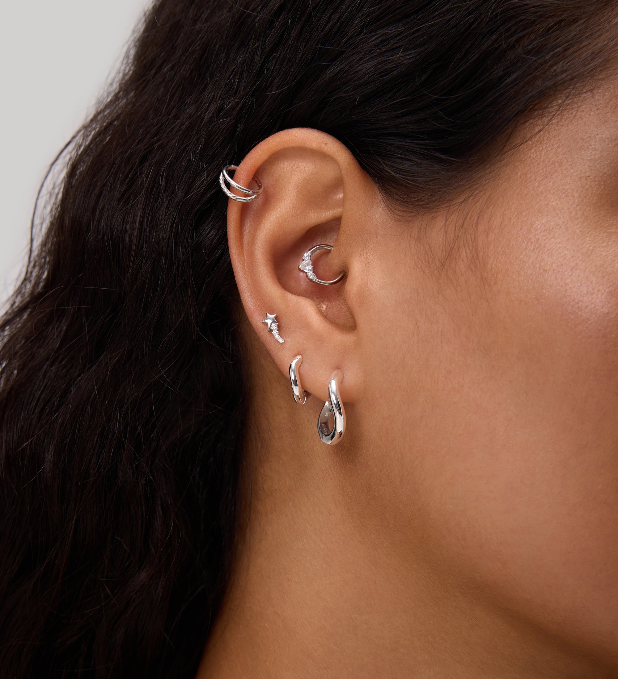 Close-up of a person's ear adorned with multiple earrings, including the Nori Twisted Hoop Earrings in Gold on the lobe. The person has long, dark hair and light brown skin.