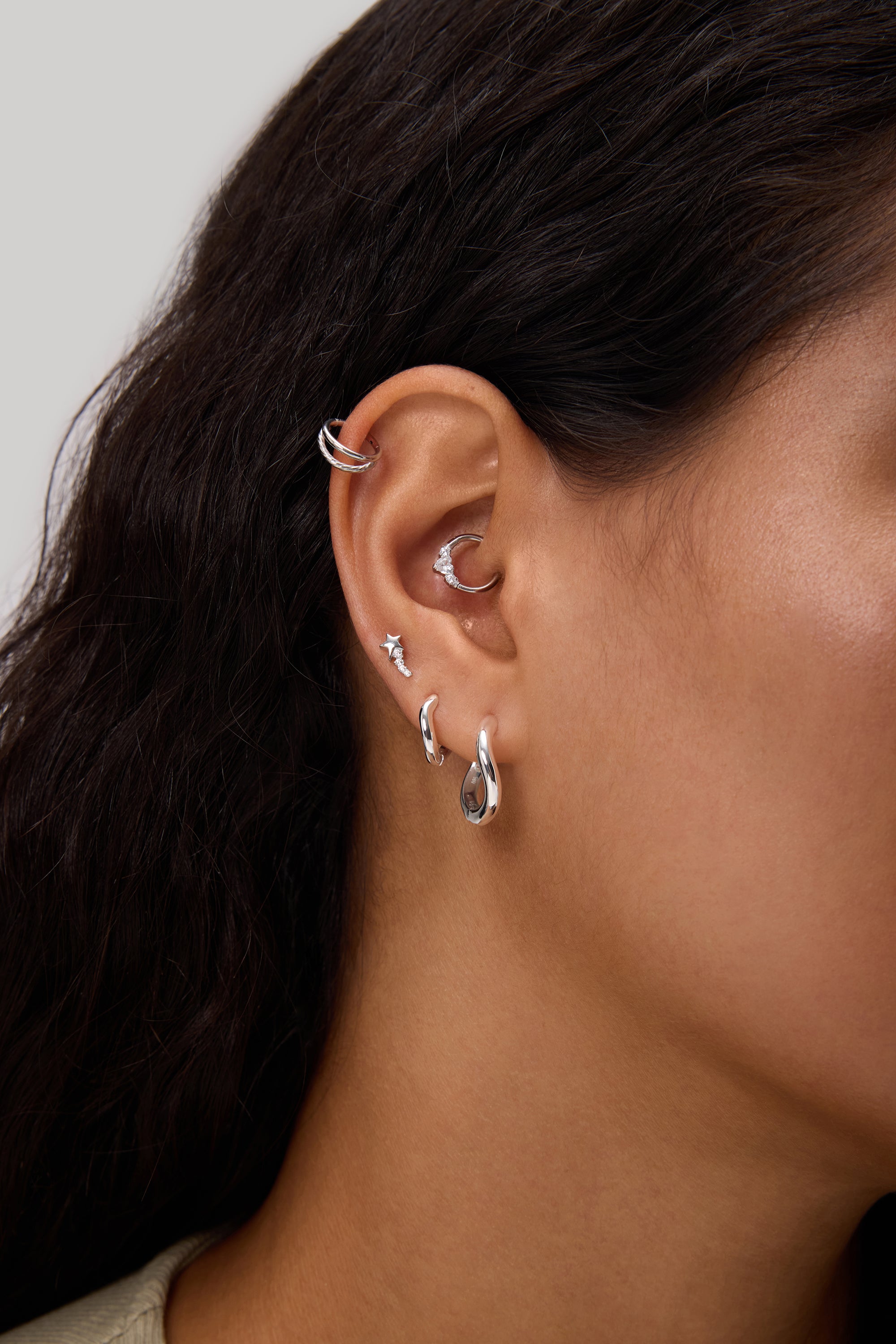 Close-up of a person's ear adorned with multiple earrings, including the Nori Twisted Hoop Earrings in Gold on the lobe. The person has long, dark hair and light brown skin.