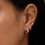 A close-up of a person's ear showcases silver and diamond earrings—including hoops, studs, a bow-shaped earring, and the Nori Twisted Crystal Huggies in Gold—adorned with sparkling cubic zirconia along the earlobe and cartilage.