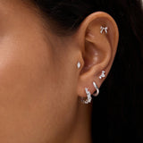 A close-up of a person's ear showcases silver and diamond earrings—including hoops, studs, a bow-shaped earring, and the Nori Twisted Crystal Huggies in Gold—adorned with sparkling cubic zirconia along the earlobe and cartilage.