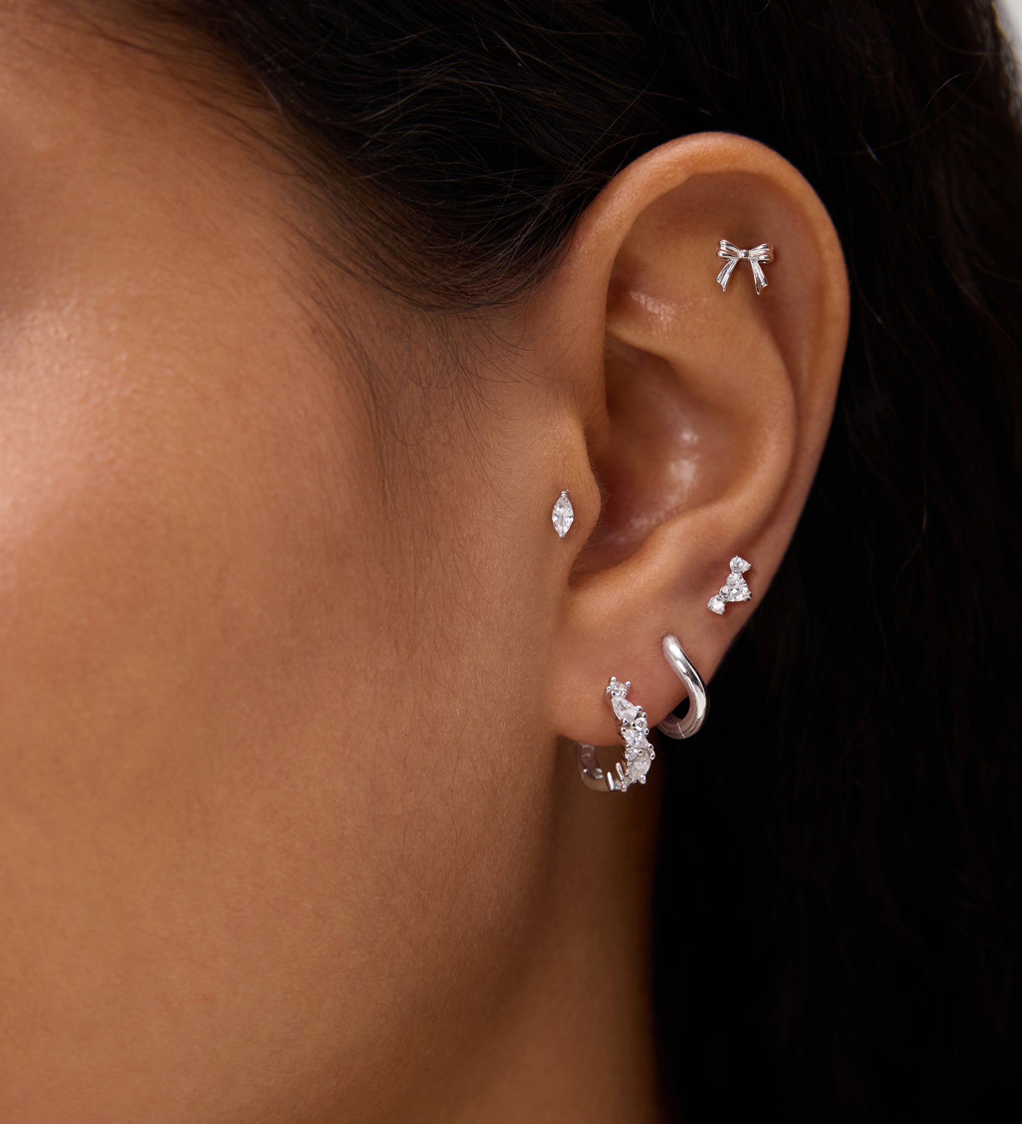 A close-up of a person's ear showcases silver and diamond earrings—including hoops, studs, a bow-shaped earring, and the Nori Twisted Crystal Huggies in Gold—adorned with sparkling cubic zirconia along the earlobe and cartilage.