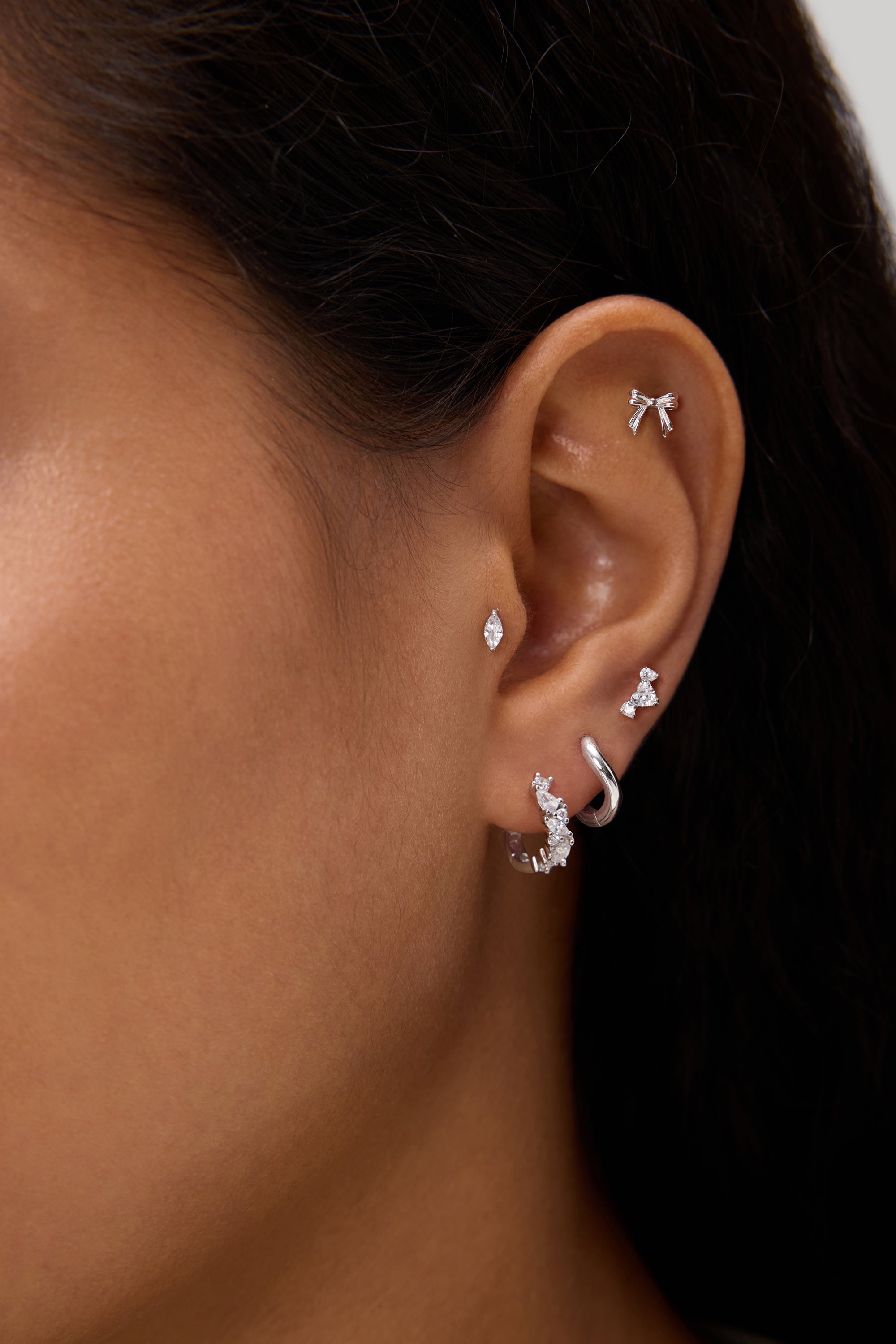 A close-up of a person's ear showcases silver and diamond earrings—including hoops, studs, a bow-shaped earring, and the Nori Twisted Crystal Huggies in Gold—adorned with sparkling cubic zirconia along the earlobe and cartilage.