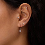 A close-up of a woman's ear adorned with five earrings, including the Flora Crystal Charm Huggies in Gold—a cubic zirconia hoop with a star charm—plus two lobe studs and three studs on the upper ear and cartilage.