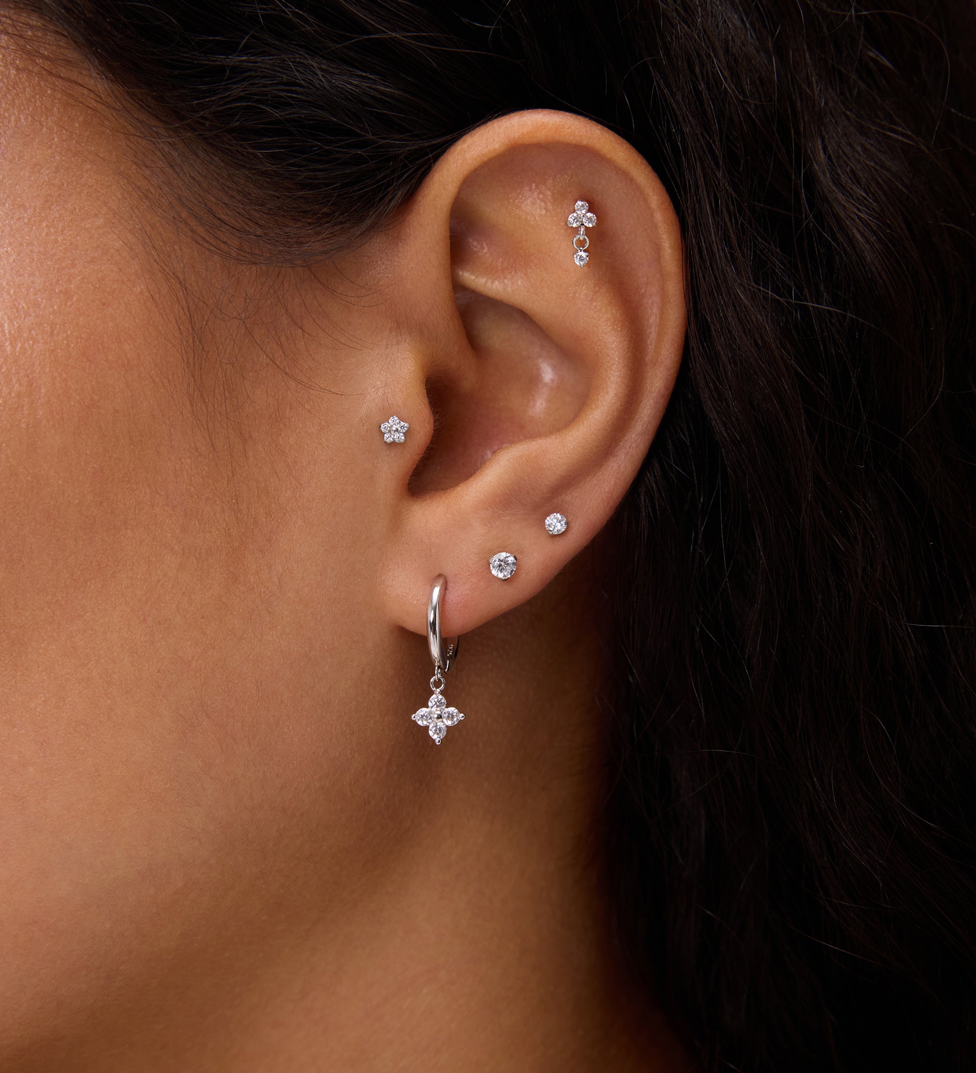 A close-up of a woman's ear adorned with five earrings, including the Flora Crystal Charm Huggies in Gold—a cubic zirconia hoop with a star charm—plus two lobe studs and three studs on the upper ear and cartilage.