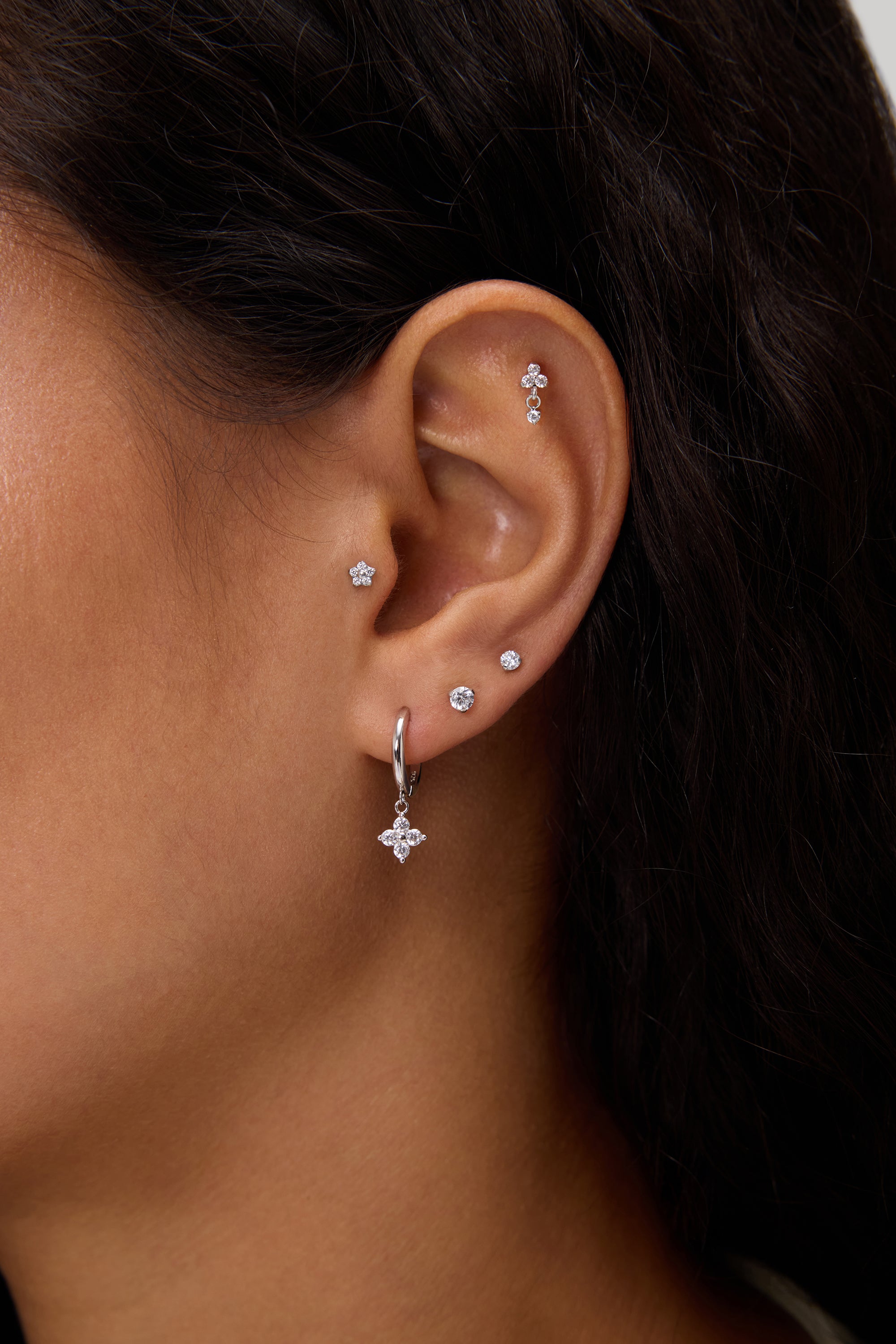 A close-up of a woman's ear adorned with five earrings, including the Flora Crystal Charm Huggies in Gold—a cubic zirconia hoop with a star charm—plus two lobe studs and three studs on the upper ear and cartilage.