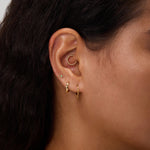 Close-up of a woman's ear with multiple gold earrings, including small hoops, a curved beaded cuff, and a 14K Solid White Gold Lab-Grown Diamond Evil Eye Flat Back Stud. She has dark wavy hair and light brown skin against a neutral background.
