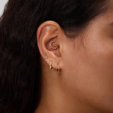 Close-up of a woman's ear with multiple gold earrings, including small hoops, a curved beaded cuff, and a 14K Solid White Gold Lab-Grown Diamond Evil Eye Flat Back Stud. She has dark wavy hair and light brown skin against a neutral background.