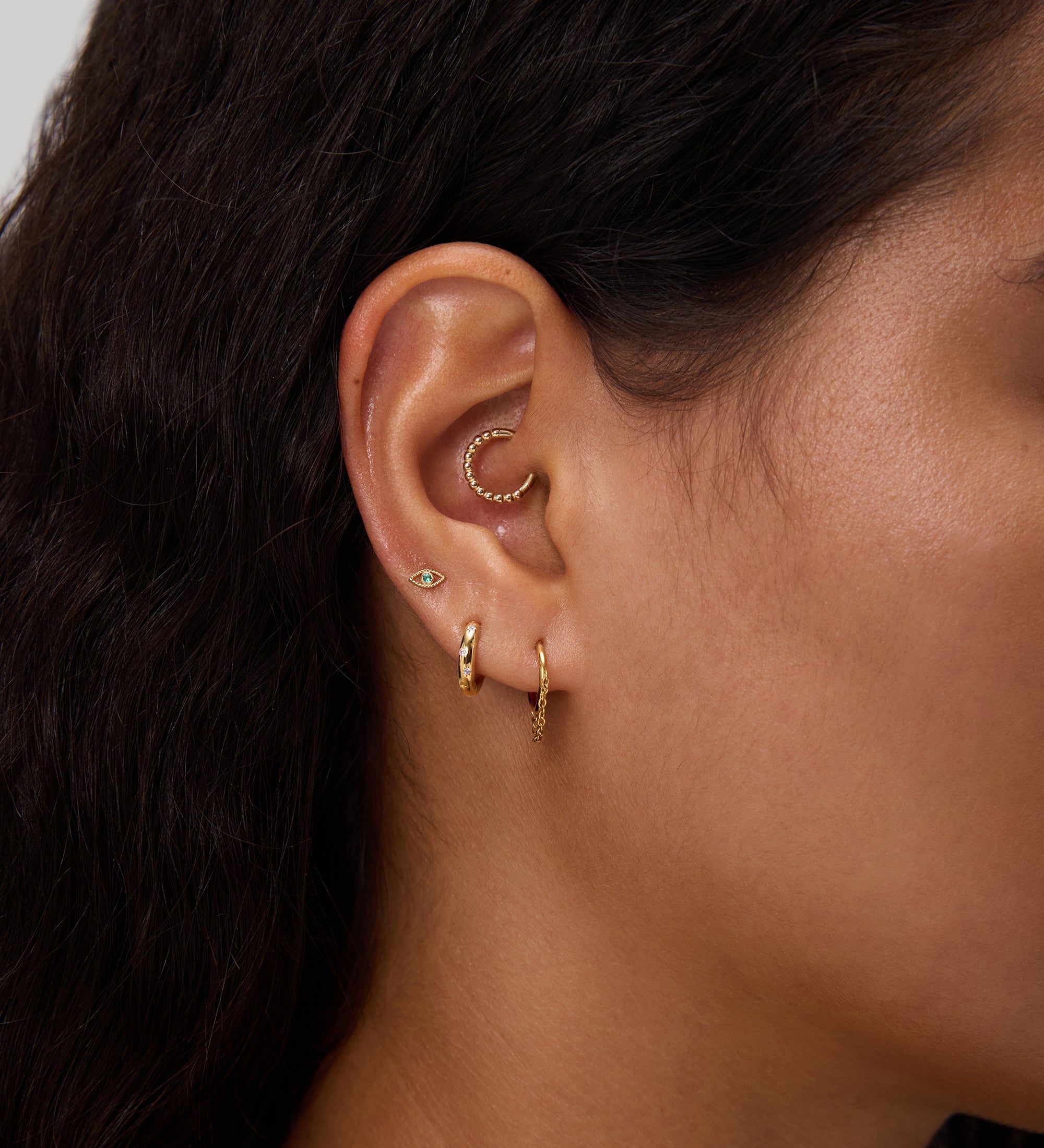 Close-up of a woman's ear with multiple gold earrings, including small hoops, a curved beaded cuff, and a 14K Solid White Gold Lab-Grown Diamond Evil Eye Flat Back Stud. She has dark wavy hair and light brown skin against a neutral background.