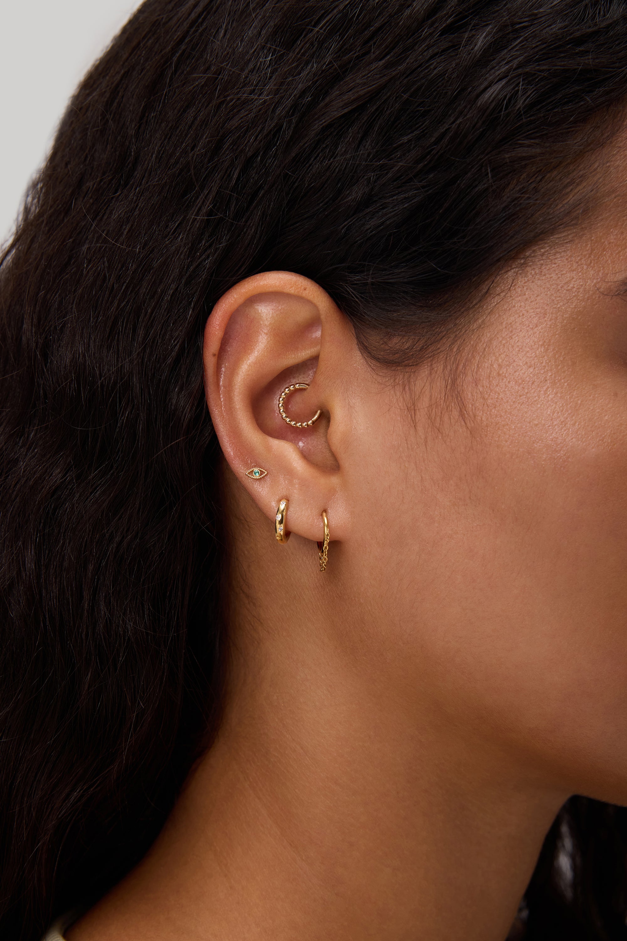 A close-up of a woman's ear adorned with multiple gold earrings—including the elegant Starfall Huggies in Gold, 14K gold plated hoops, a cartilage cuff, and sparkling cubic zirconia styles—set against her dark wavy hair and brown skin.