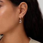 Close-up of a woman's ear adorned with the Crescent Moon Crystal Charm Hoop in Silver, alongside several small studs and hoops. She has long dark hair and is wearing a beige sleeveless top.
