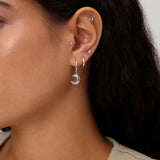 Close-up of a woman's ear adorned with the Crescent Moon Crystal Charm Hoop in Silver, alongside several small studs and hoops. She has long dark hair and is wearing a beige sleeveless top.