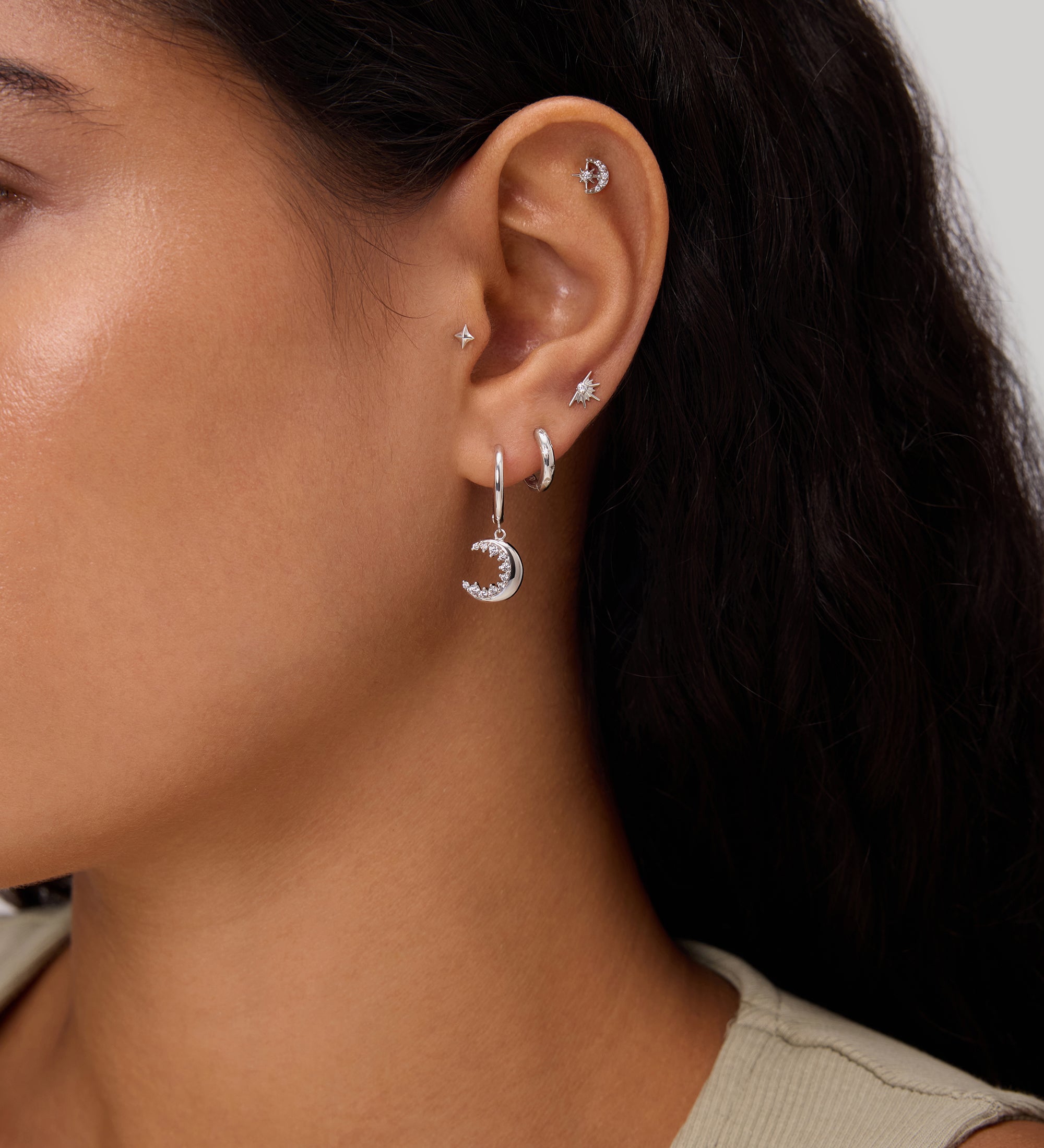 Close-up of a woman's ear adorned with the Crescent Moon Crystal Charm Hoop in Silver, alongside several small studs and hoops. She has long dark hair and is wearing a beige sleeveless top.