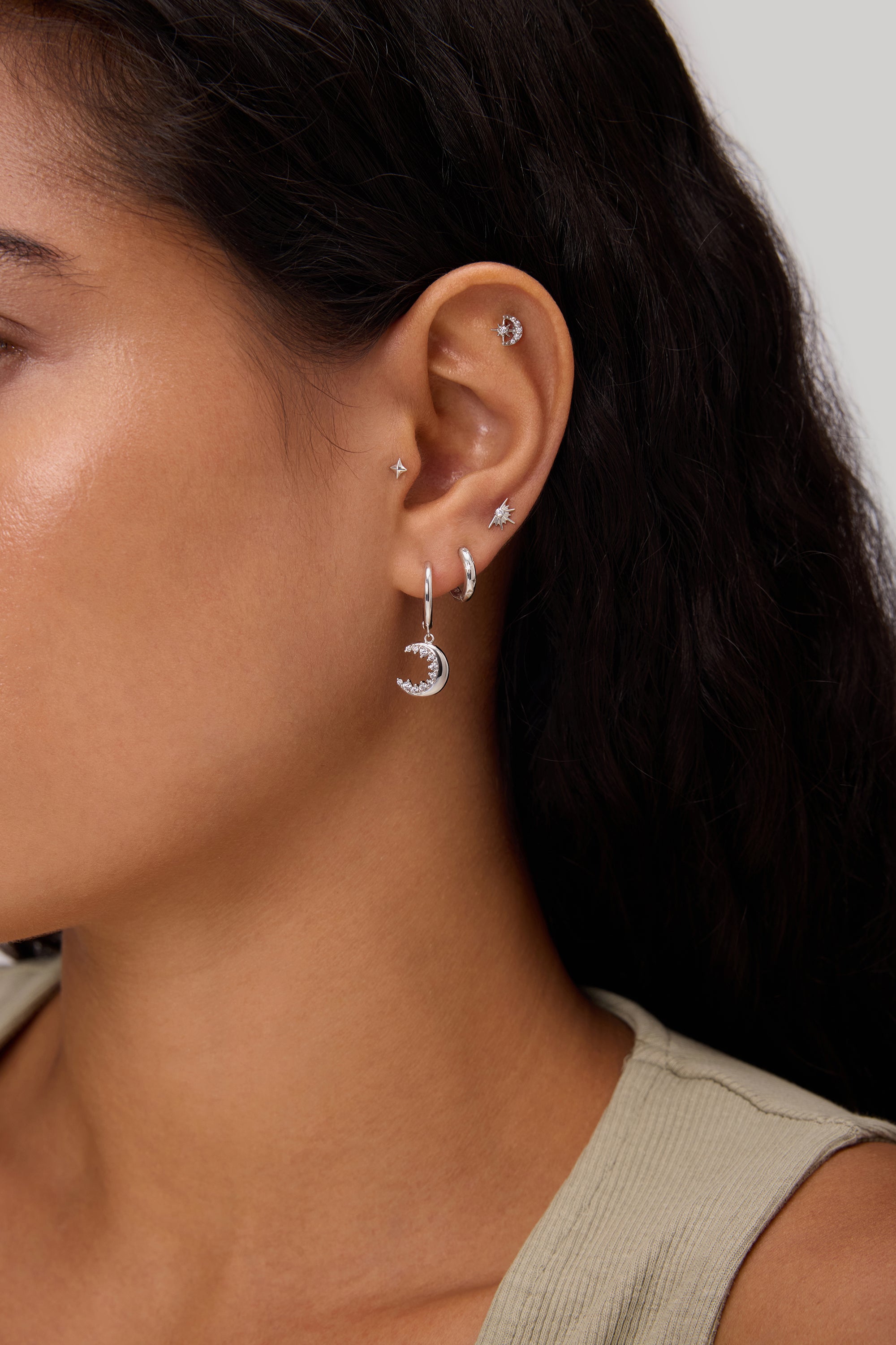 Close-up of a woman's ear adorned with the Crescent Moon Crystal Charm Hoop in Silver, alongside several small studs and hoops. She has long dark hair and is wearing a beige sleeveless top.