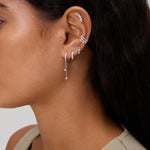 A close-up of a woman's ear adorned with the Star Double Chain Dangle Huggies in Gold, styled among other earrings, set against a neutral background. She has long dark hair and is wearing a light-colored top.