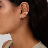 A close-up of a woman's ear adorned with the Star Double Chain Dangle Huggies in Gold, styled among other earrings, set against a neutral background. She has long dark hair and is wearing a light-colored top.