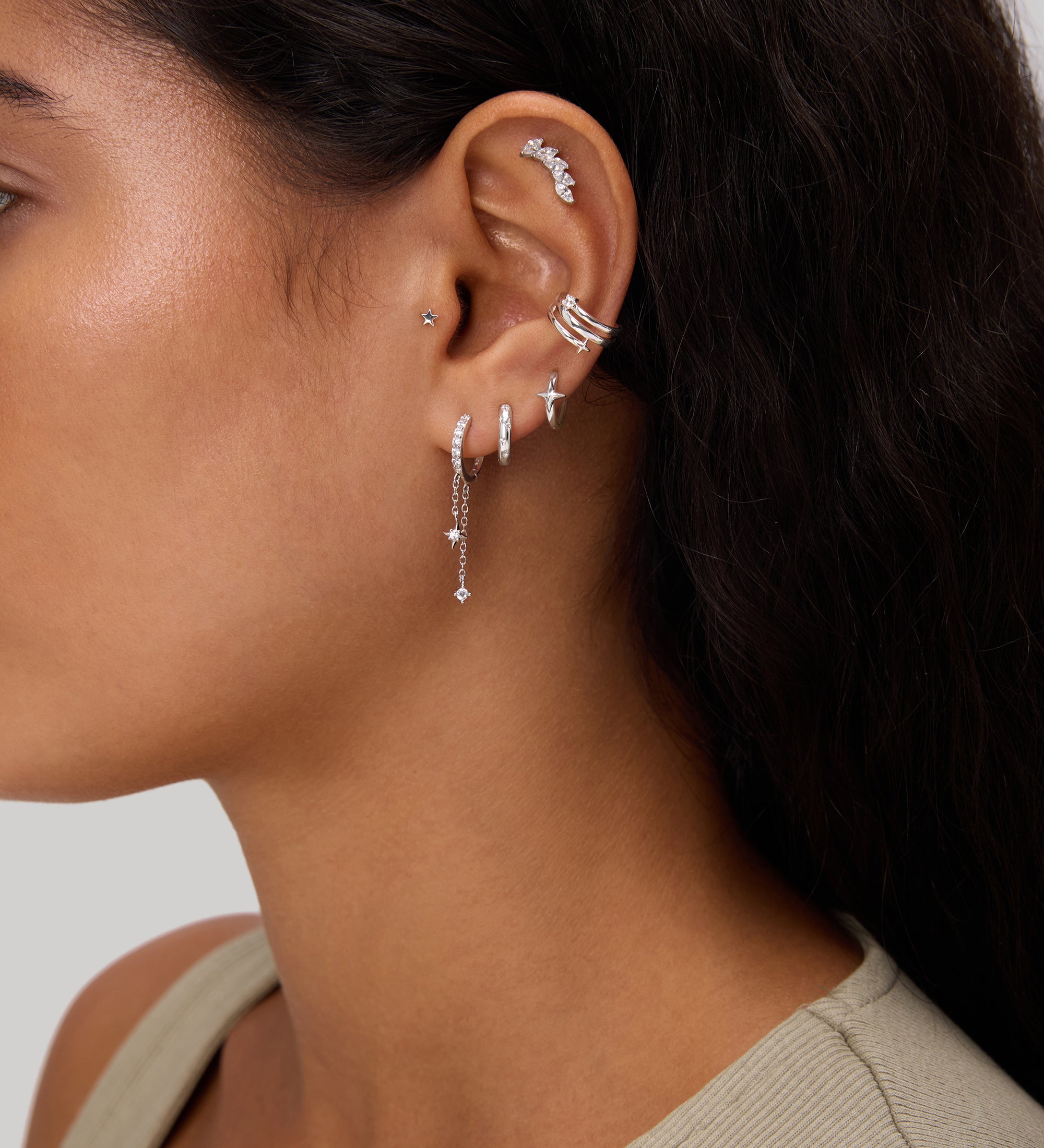 A close-up of a woman's ear adorned with the Star Double Chain Dangle Huggies in Gold, styled among other earrings, set against a neutral background. She has long dark hair and is wearing a light-colored top.