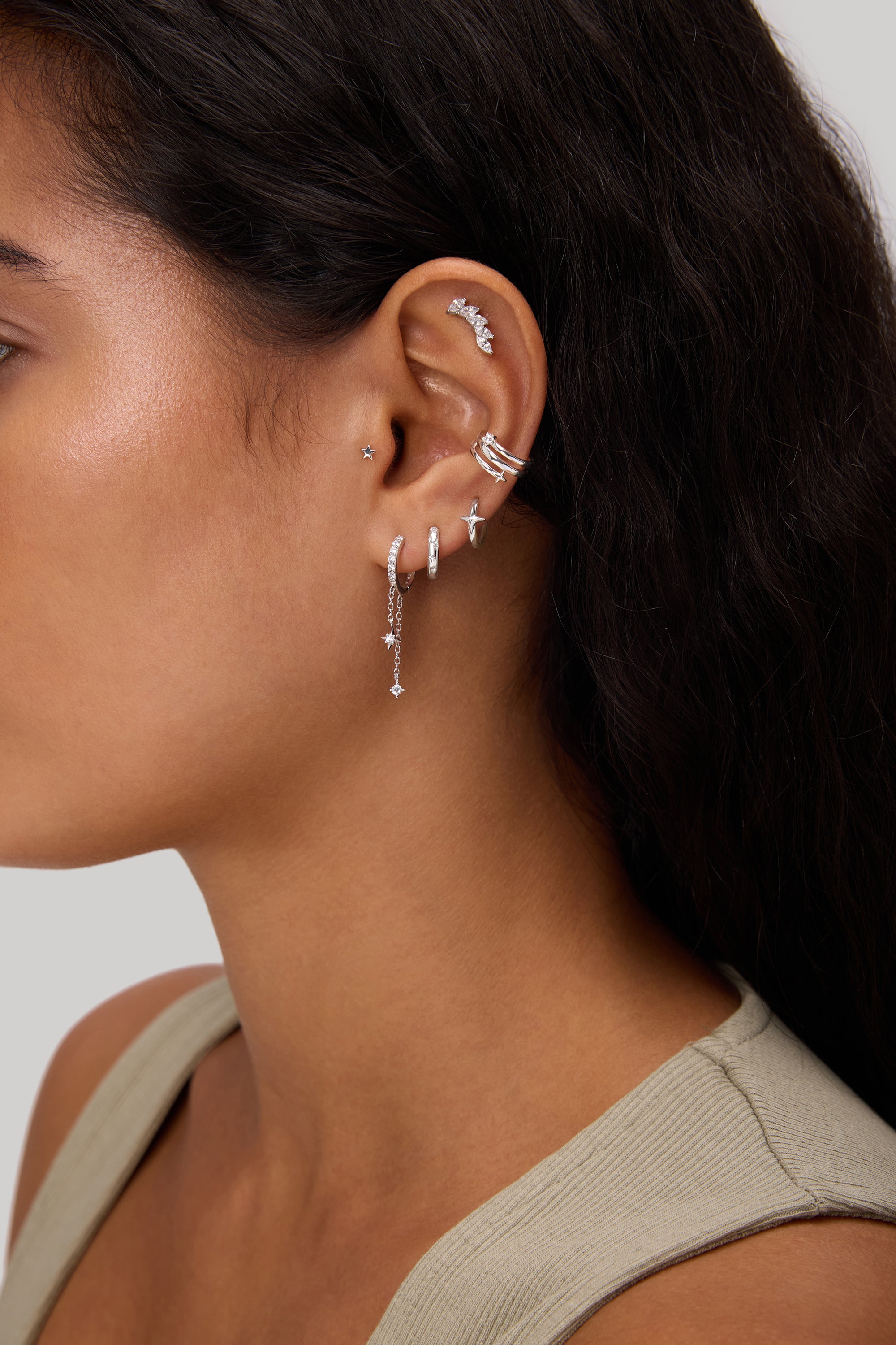 A close-up of a woman's ear adorned with the Star Double Chain Dangle Huggies in Gold, styled among other earrings, set against a neutral background. She has long dark hair and is wearing a light-colored top.
