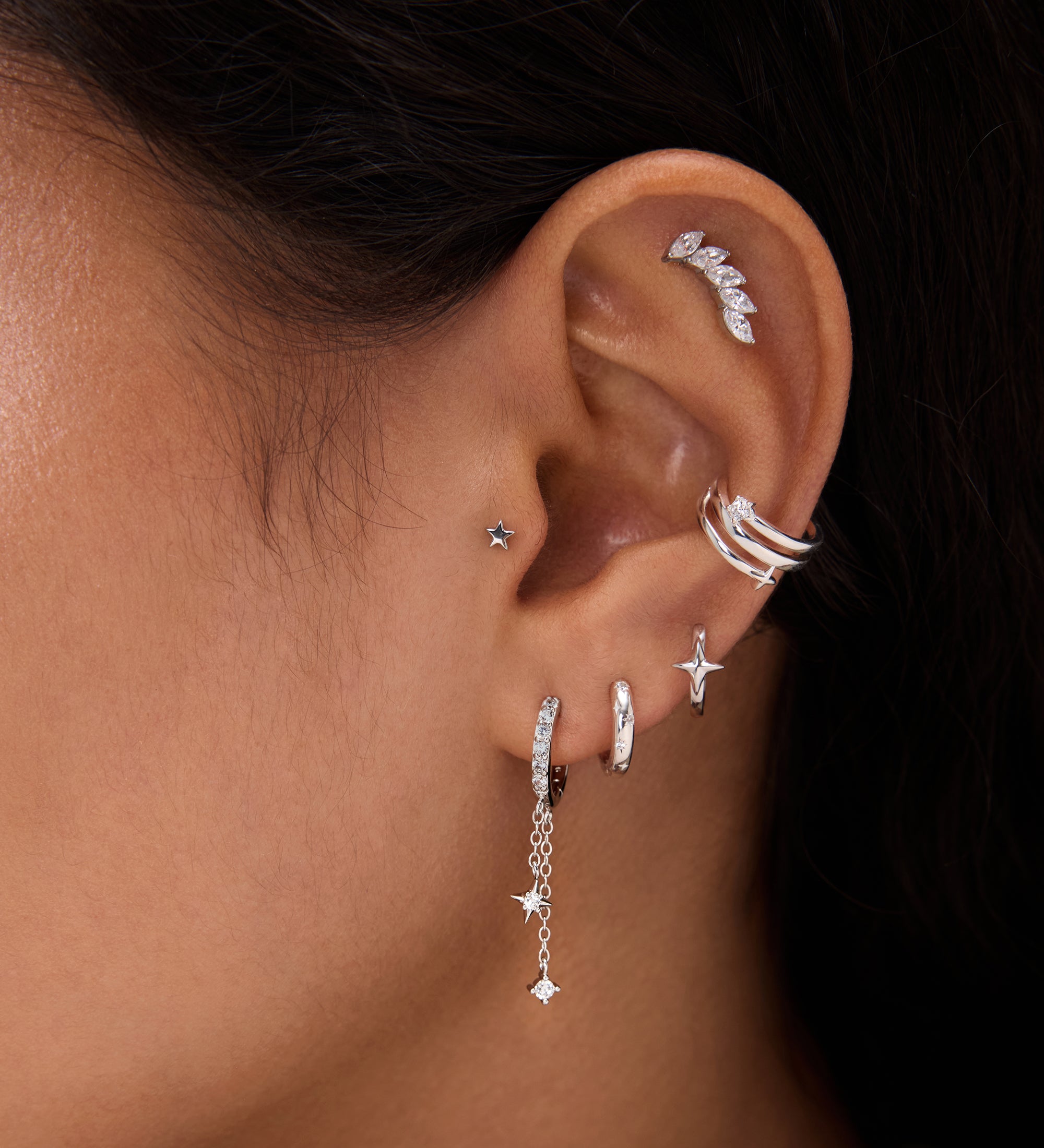 Close-up of a person with dark hair and warm skin tone wearing multiple silver and diamond earrings, including the 14K Solid White Gold Mini Star Flat Back Stud, hoops, dangling chains, and an ear cuff.
