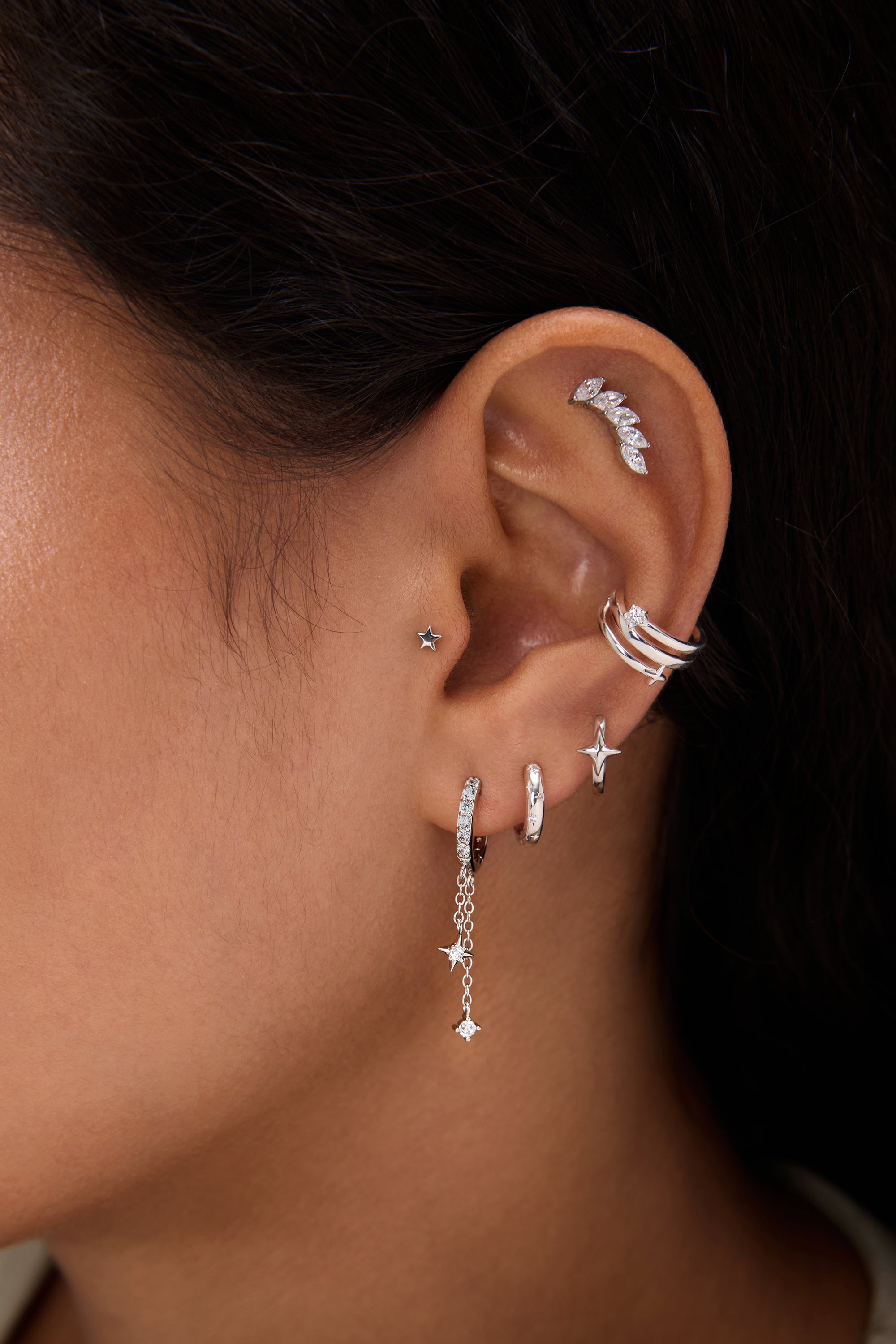 Close-up of a person with dark hair and warm skin tone wearing multiple silver and diamond earrings, including the 14K Solid White Gold Mini Star Flat Back Stud, hoops, dangling chains, and an ear cuff.
