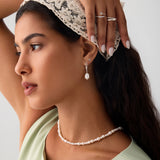 A woman with long dark hair wears a lace headscarf, pearl jewelry, and a light green top. She highlights her helix piercing adorned with the 14K Solid White Gold Shell Flat Back Stud while posing with one arm raised against a plain background.