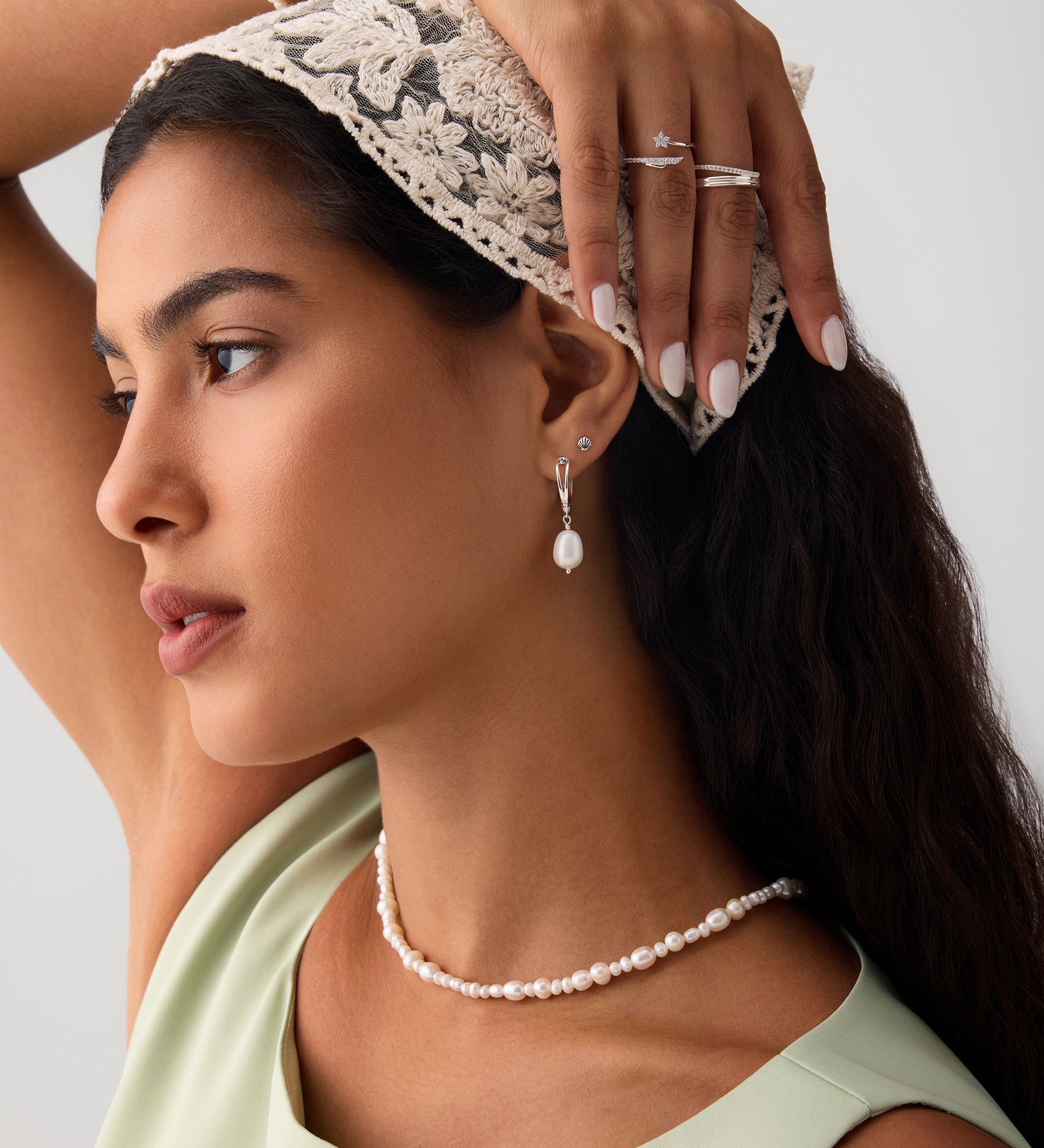 A woman with long dark hair wears a lace headscarf, pearl jewelry, and a light green top. She highlights her helix piercing adorned with the 14K Solid White Gold Shell Flat Back Stud while posing with one arm raised against a plain background.