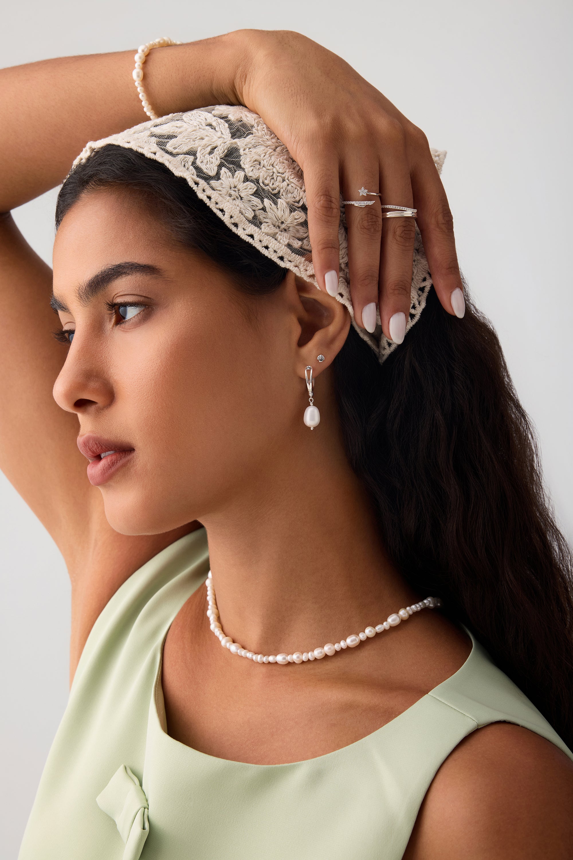 A woman with long dark hair wears a lace headscarf, pearl jewelry, and a light green top. She highlights her helix piercing adorned with the 14K Solid White Gold Shell Flat Back Stud while posing with one arm raised against a plain background.