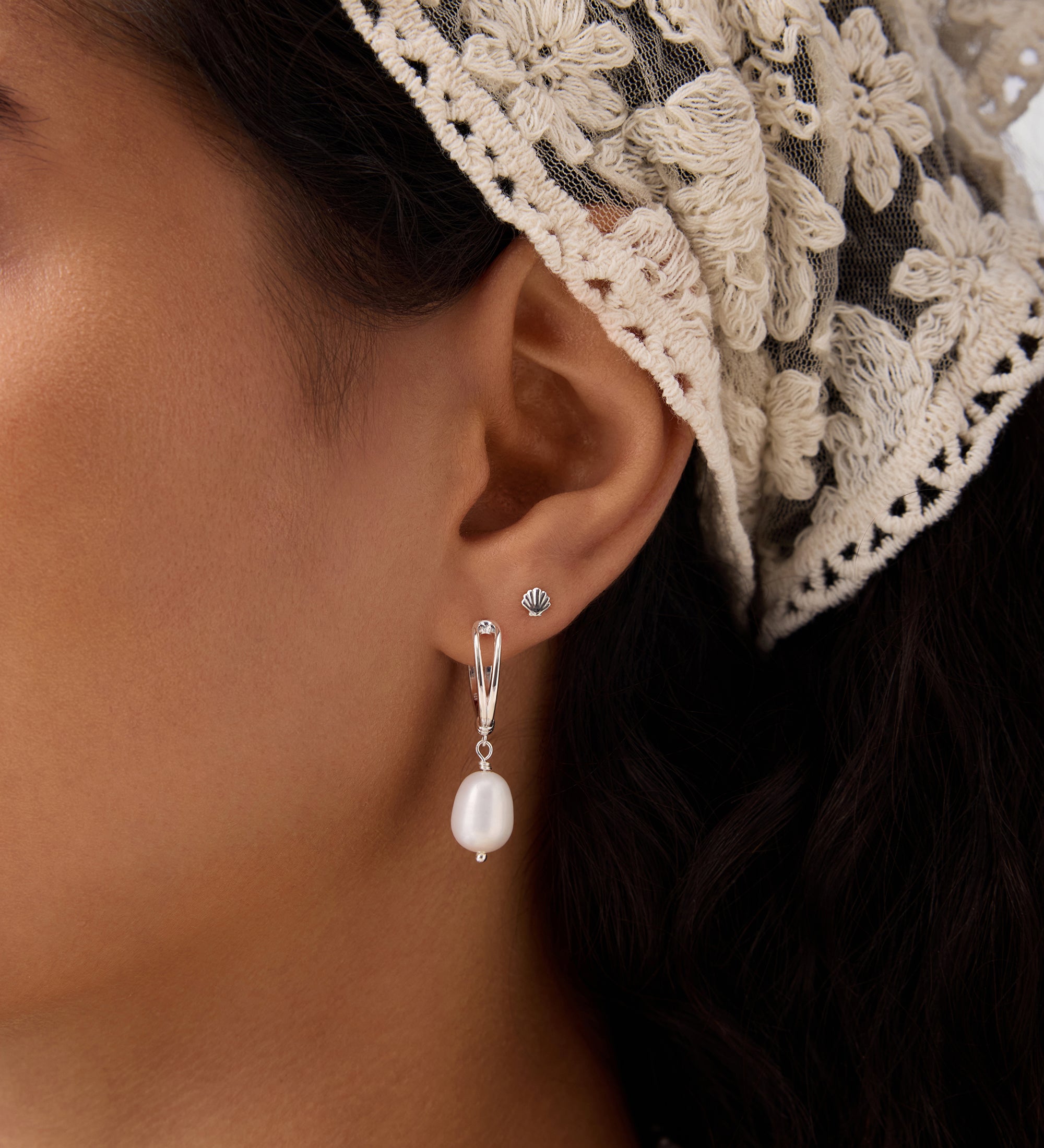 Close-up of a woman's ear wearing the 14K Solid Yellow Gold Shell Flat Back Stud and a silver drop earring with a pearl. She has dark hair, a cream lace headscarf, and a matching pearl necklace partially visible.