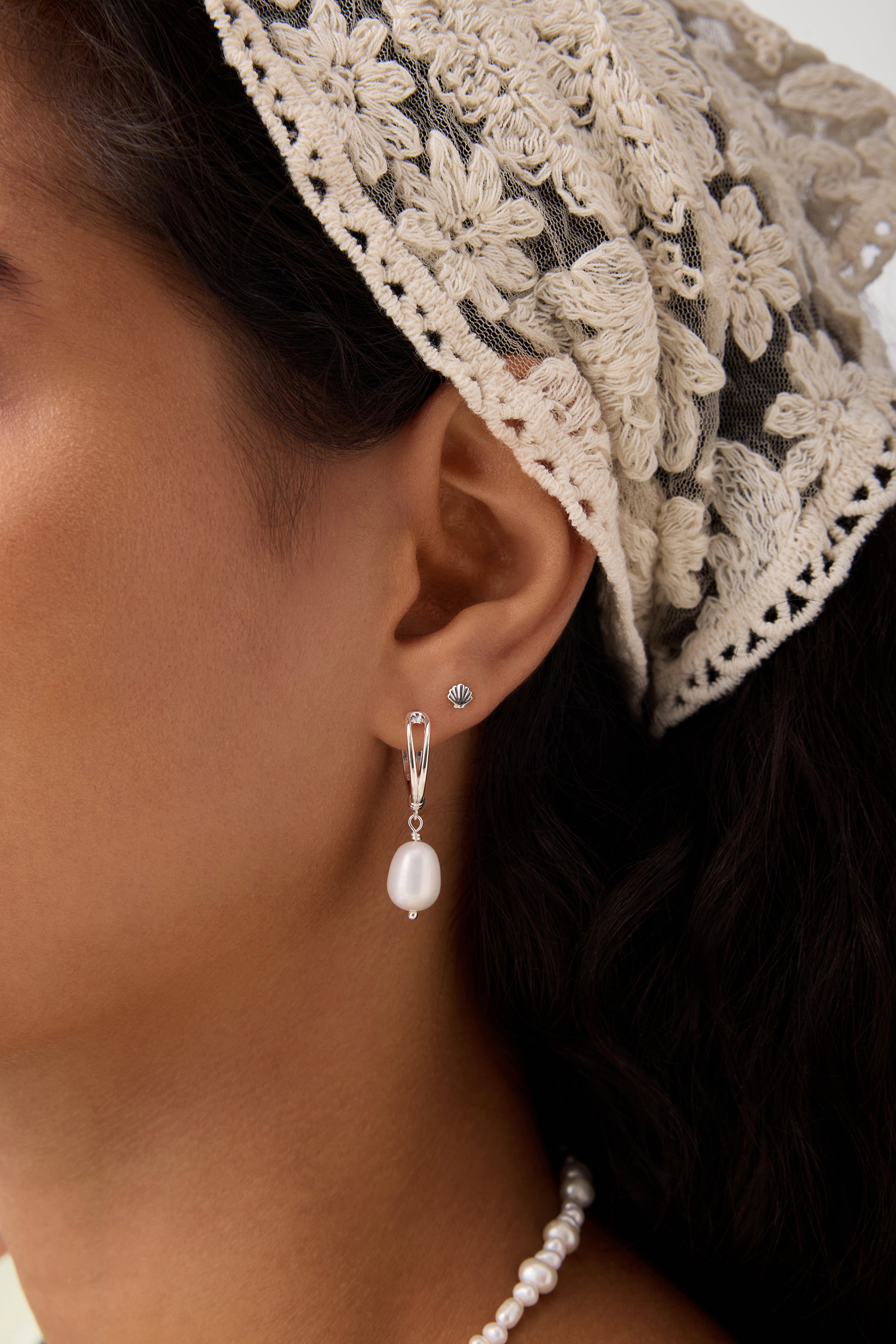 Close-up of a woman's ear wearing the 14K Solid Yellow Gold Shell Flat Back Stud and a silver drop earring with a pearl. She has dark hair, a cream lace headscarf, and a matching pearl necklace partially visible.