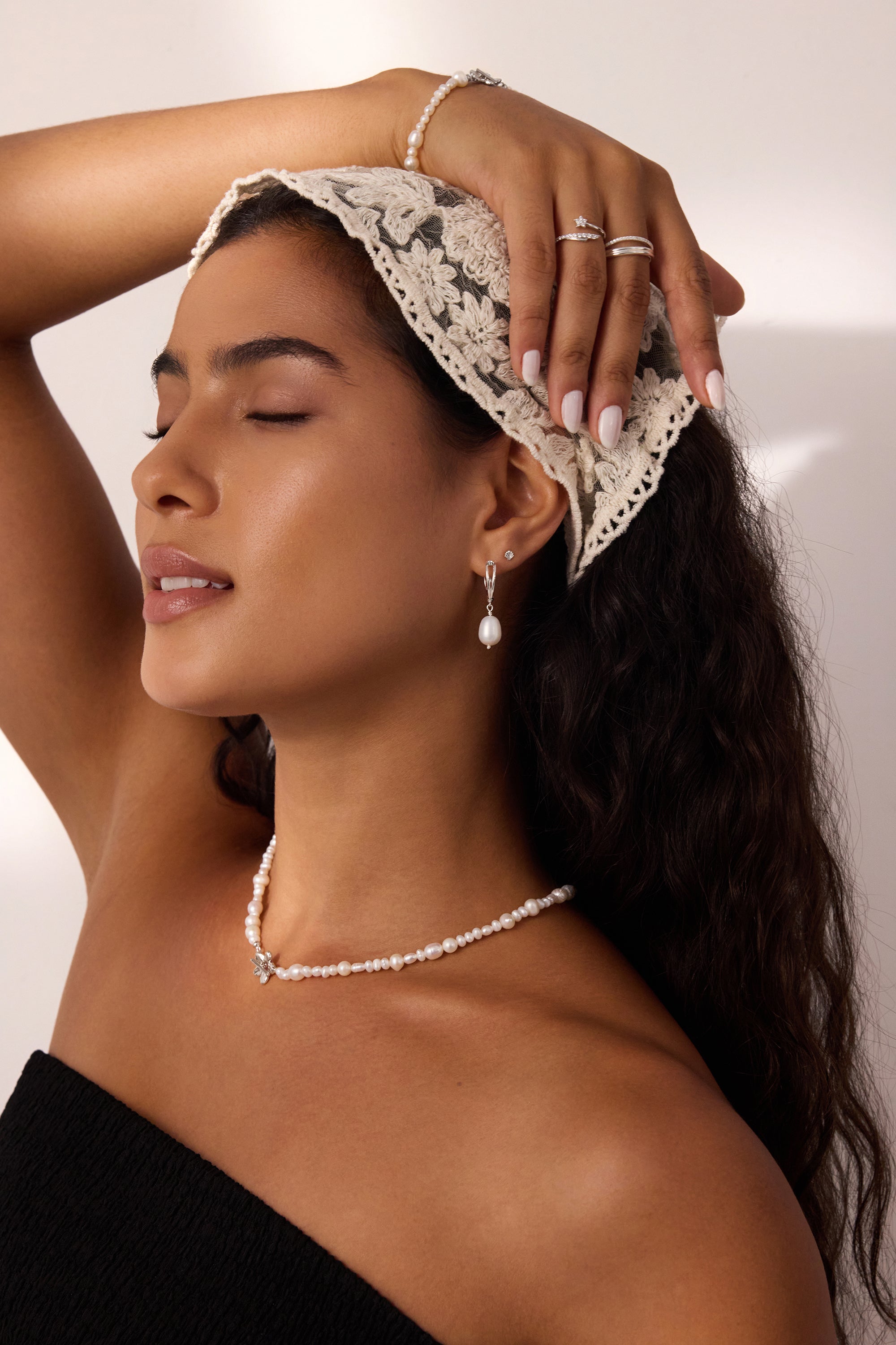 A woman with long dark hair, wearing a lace headscarf, pearl jewelry, and a black top, poses with one arm raised and eyes closed in soft lighting, showcasing the 14K Solid White Gold Shell Flat Back Stud.