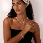 A woman with long dark hair wears a cream lace headscarf, Freshwater Pearl Charm Huggies in Silver, and a black strapless top, standing against a light background and looking to the side with a relaxed expression.