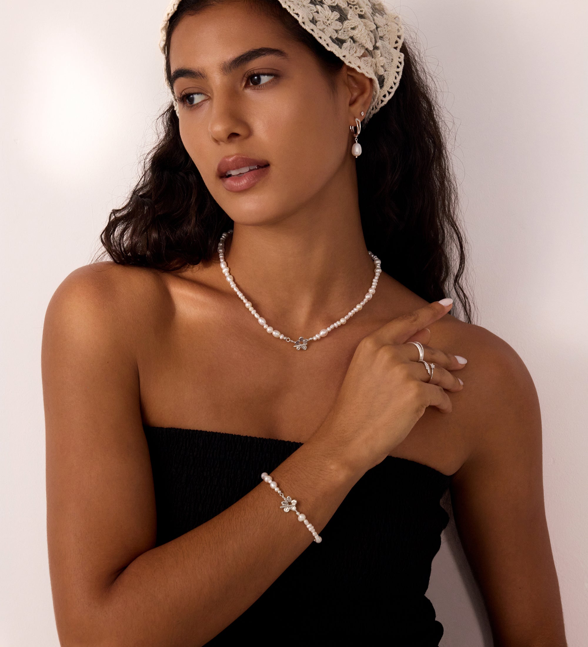 Woman wearing a cream lace headscarf, black strapless top, Freshwater Pearl Charm Hoops in Silver, and rings, standing against a white background, looking to the side with a relaxed expression.