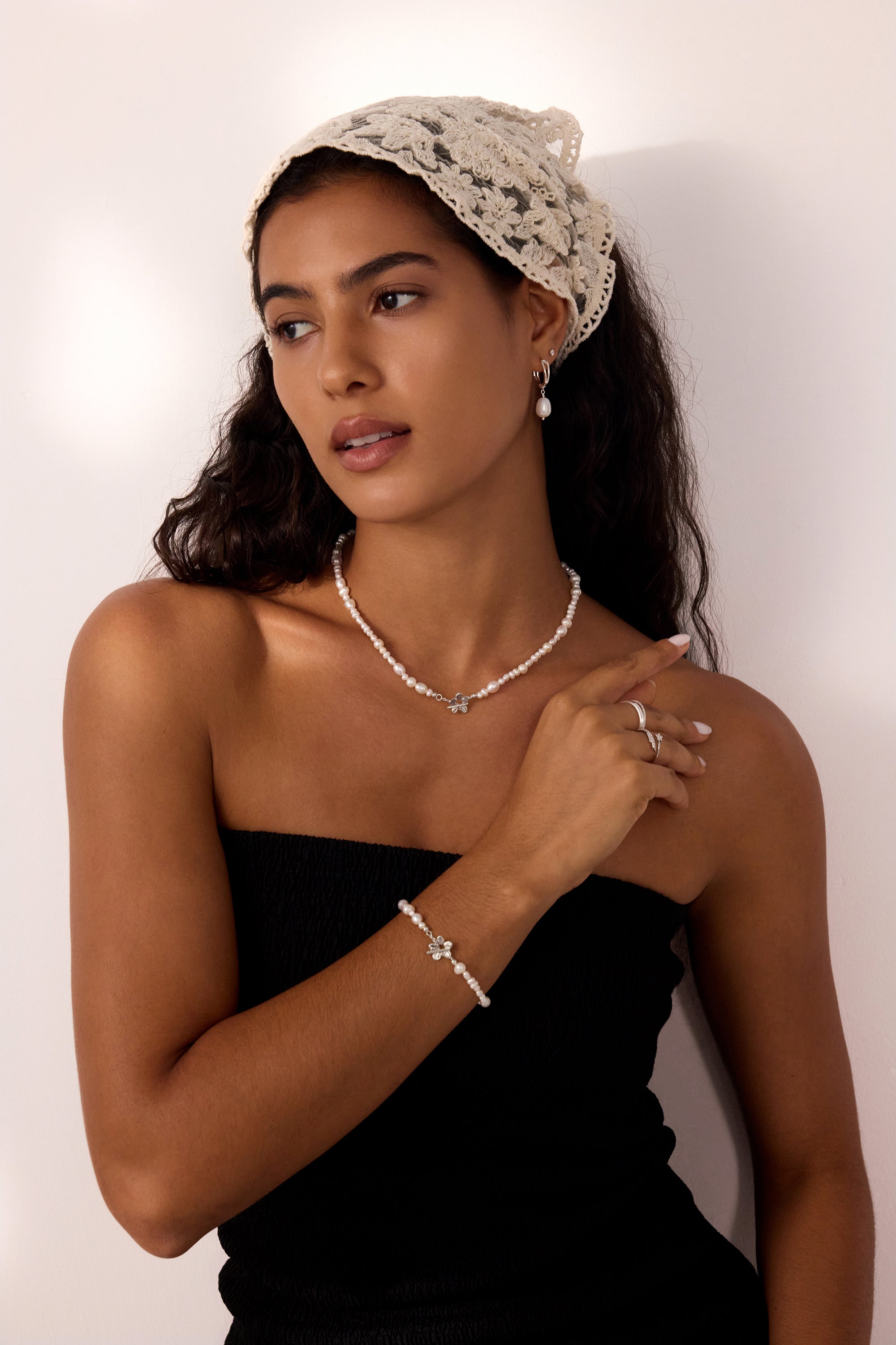 A woman with long dark hair wears a cream lace headscarf, Freshwater Pearl Charm Huggies in Silver, and a black strapless top, standing against a light background and looking to the side with a relaxed expression.