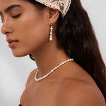A woman with long dark hair is seen in profile, eyes closed, wearing a lace headscarf and the Knot Multi Pearl Drop Earrings in Gold, crafted from freshwater pearls. She pairs them with a matching pearl necklace and a strapless black top against a plain background.