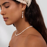 A woman with long dark hair is seen in profile, eyes closed, wearing a lace headscarf and the Knot Multi Pearl Drop Earrings in Gold, crafted from freshwater pearls. She pairs them with a matching pearl necklace and a strapless black top against a plain background.