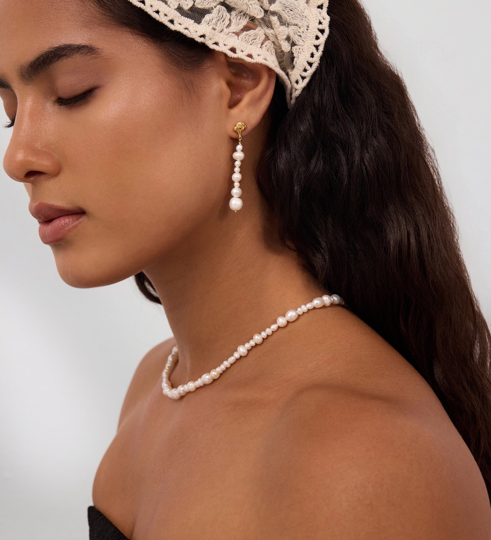 A woman with long dark hair is seen in profile, eyes closed, wearing a lace headscarf and the Knot Multi Pearl Drop Earrings in Gold, crafted from freshwater pearls. She pairs them with a matching pearl necklace and a strapless black top against a plain background.