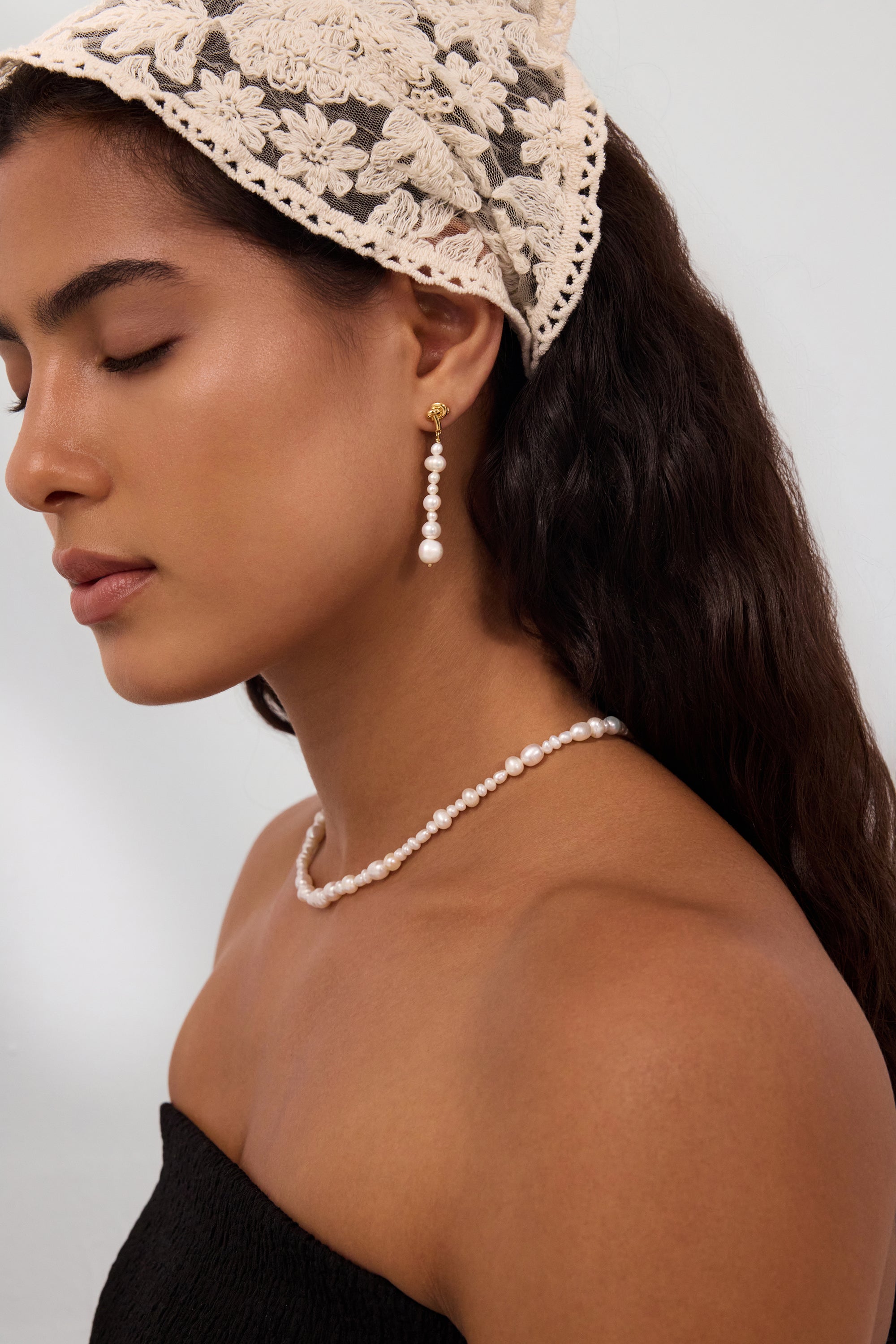 A woman with long dark hair is seen in profile, eyes closed, wearing a lace headscarf and the Knot Multi Pearl Drop Earrings in Gold, crafted from freshwater pearls. She pairs them with a matching pearl necklace and a strapless black top against a plain background.