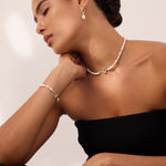 Woman with dark hair in a low bun wearing a strapless black top, pearl necklace, matching bracelet, and Freshwater Pearl Charm Huggies in Silver—resting her chin on her hand with eyes closed against a neutral background.