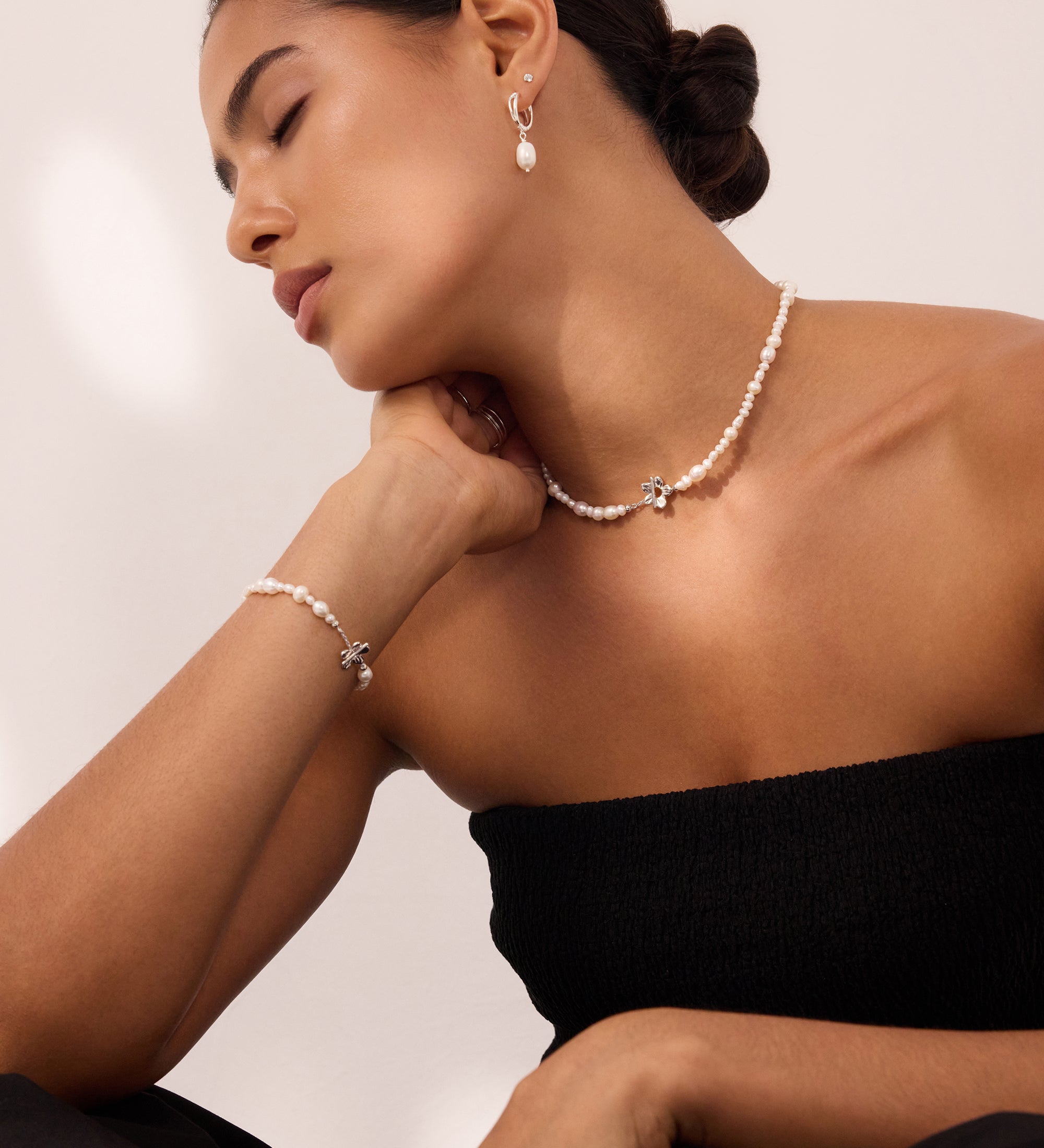 Woman with dark hair in a low bun wearing a strapless black top, pearl necklace, matching bracelet, and Freshwater Pearl Charm Huggies in Silver—resting her chin on her hand with eyes closed against a neutral background.