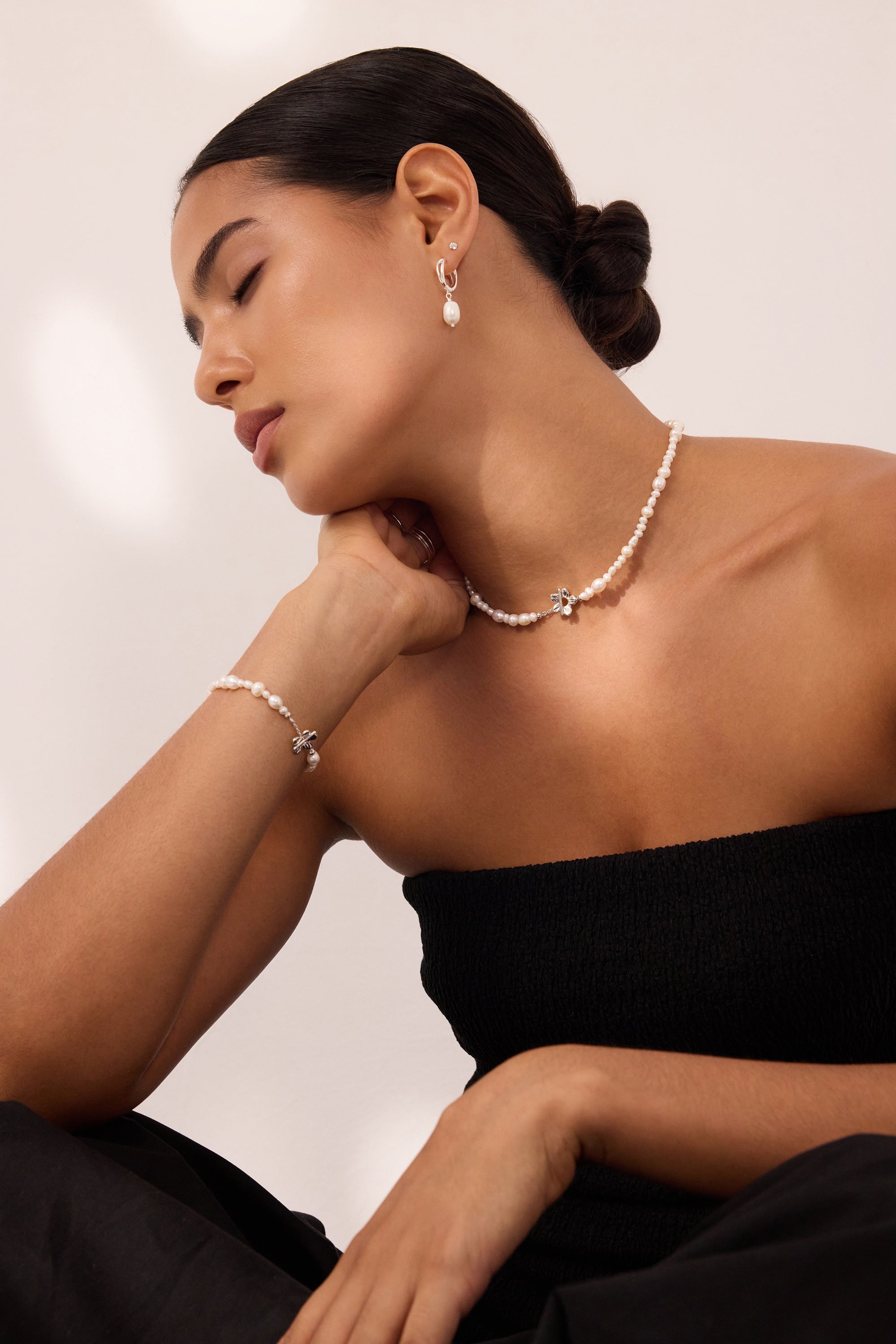 Woman with dark hair in a low bun wearing a strapless black top, pearl necklace, matching bracelet, and Freshwater Pearl Charm Huggies in Silver—resting her chin on her hand with eyes closed against a neutral background.