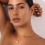 A woman with tanned skin and dark hair pulled back poses with one arm raised, wearing a black textured top, matching pearl drop earrings, and a gold Mini Paperclip Double Chain Necklace in Gold with a pearl pendant.