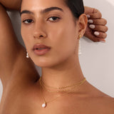 A woman with tanned skin and dark hair pulled back poses with one arm raised, wearing a black textured top, matching pearl drop earrings, and a gold Mini Paperclip Double Chain Necklace in Gold with a pearl pendant.