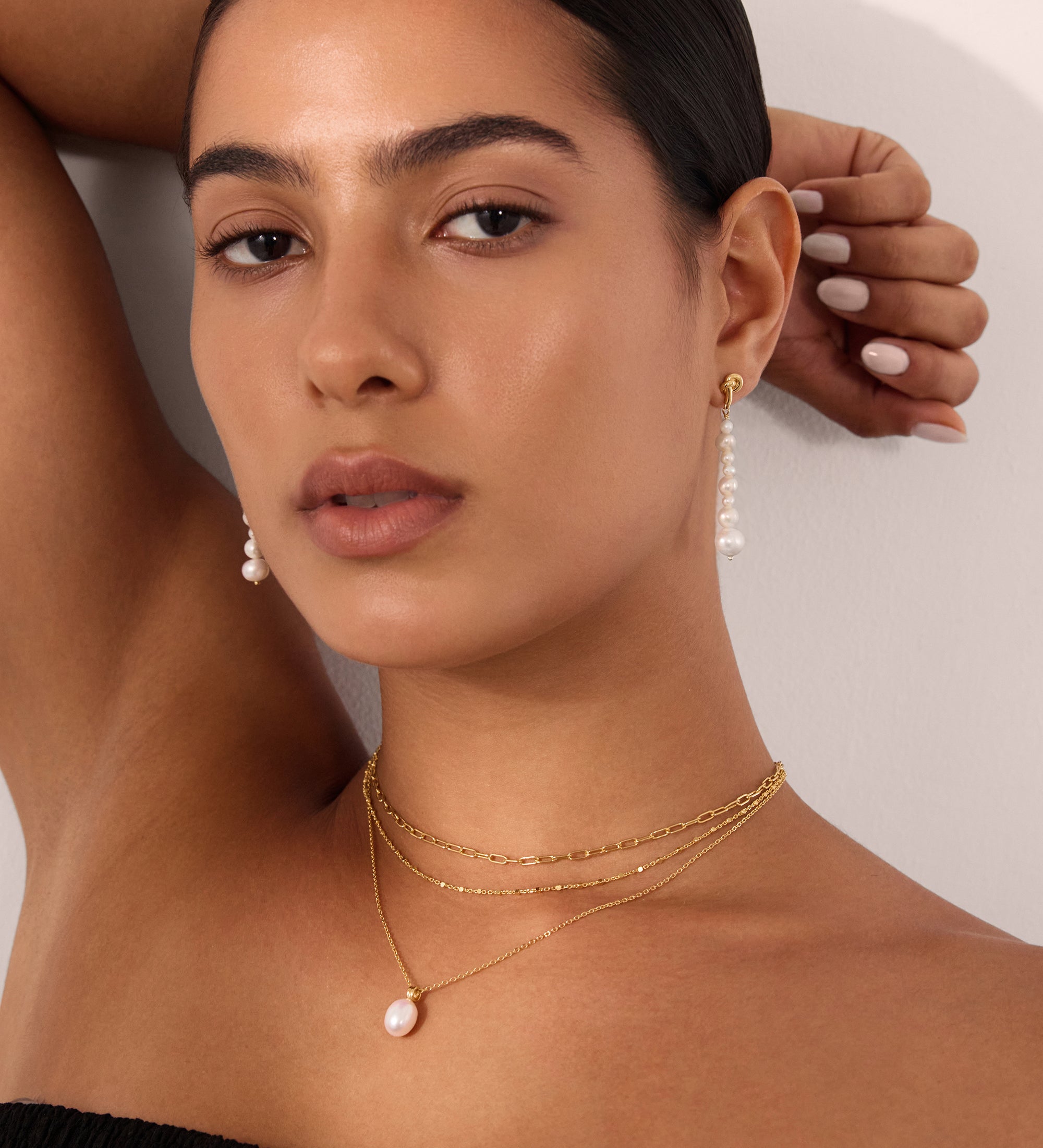 A woman with tanned skin and dark hair pulled back poses with one arm raised, wearing a black textured top, matching pearl drop earrings, and a gold Mini Paperclip Double Chain Necklace in Gold with a pearl pendant.