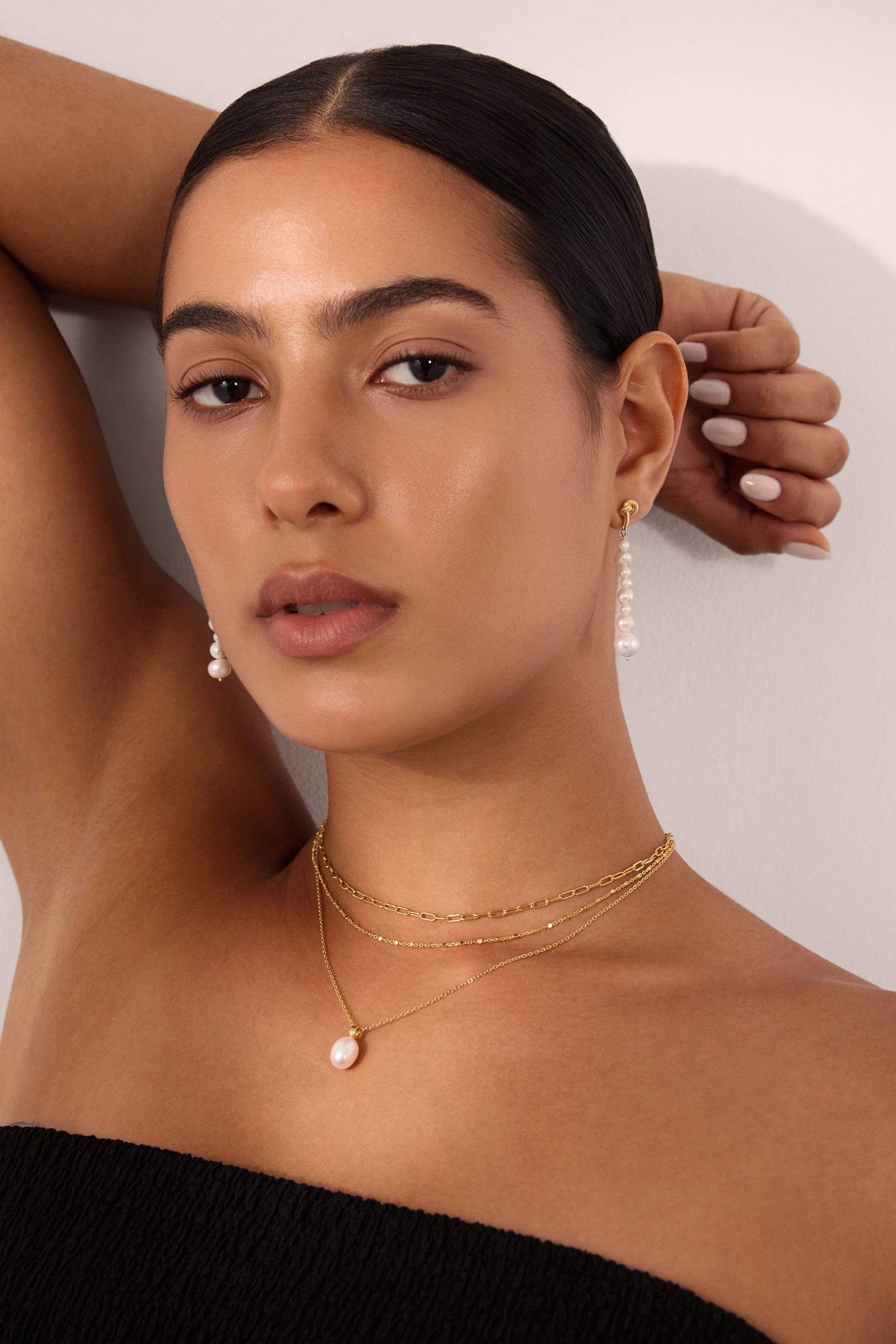 A woman with tanned skin and dark hair pulled back poses with one arm raised, wearing a black textured top, matching pearl drop earrings, and a gold Mini Paperclip Double Chain Necklace in Gold with a pearl pendant.