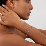 A woman with smooth skin wears Starfall Huggies in Silver, thin layered silver bracelets, and a silver ring. She rests her manicured hand on her neck, posing against a plain light background.