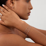 A woman with smooth skin wears Starfall Huggies in Silver, thin layered silver bracelets, and a silver ring. She rests her manicured hand on her neck, posing against a plain light background.