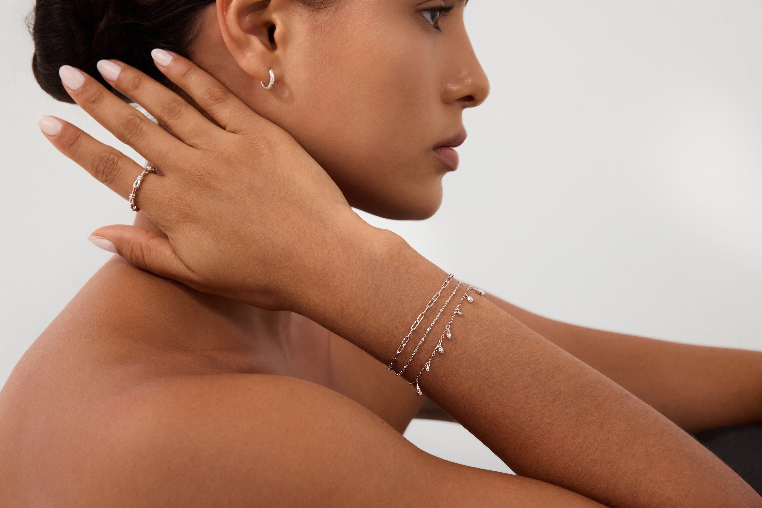 A woman with smooth skin wears Starfall Huggies in Silver, thin layered silver bracelets, and a silver ring. She rests her manicured hand on her neck, posing against a plain light background.