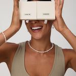 A smiling woman holds two beige VANCHI boxes over her eyes like glasses. She wears a taupe tank top, the Flora Multi Pearl Toggle Choker in Gold, along with a matching bracelet and pearl earrings against a plain background.